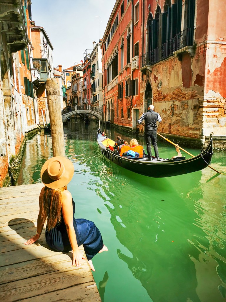 Kobieta w kapeluszu siedzi na drewnianym pomoście, obserwując gondolę płynącą wąskim zielonym kanałem w Wenecji, otoczoną kolorowymi budynkami.