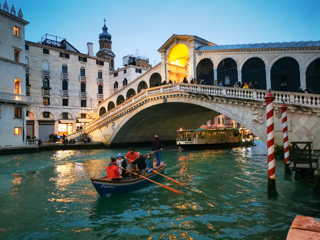 Zmierzch pod Mostem Rialto w Wenecji: niebieska łódź z wioślarzami w pomarańczowych kurtkach na zielonym kanale, z oświetlonymi arkadami.