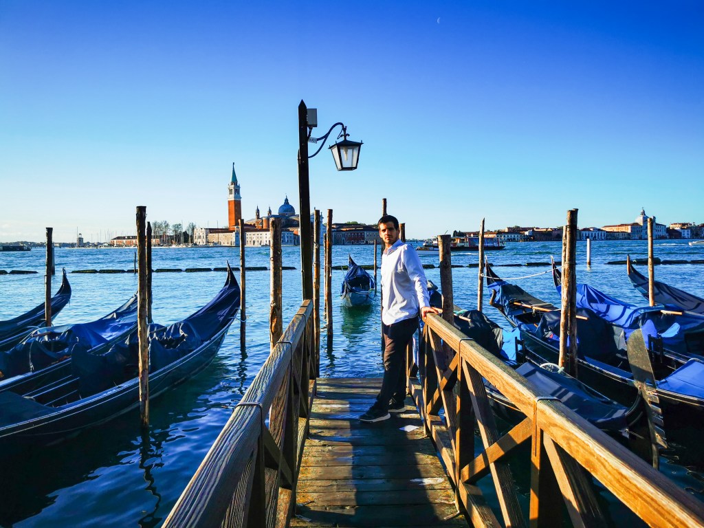 Veneza: Tudo o que deves saber antes da tua viagem Homem apoiado num cais de gôndolas com a ilha de San Giorgio Maggiore ao fundo em Veneza