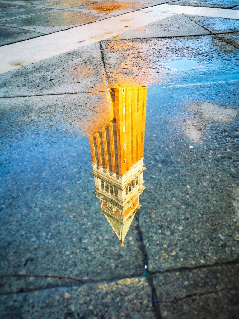 Veneza: Tudo o que deves saber antes da tua viagem Reflexo do Campanário de São Marcos numa poça sobre o pavimento em Veneza