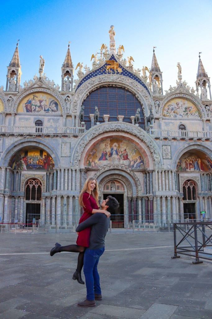 Veneza: Tudo o que deves saber antes da tua viagem Homem levanta mulher de vestido vermelho em frente à Basílica de São Marcos em Veneza