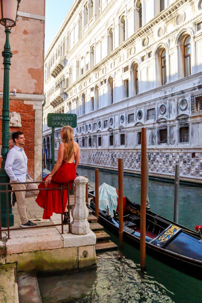 Veneza: Tudo o que deves saber antes da tua viagem Casal junto a uma gôndola em Veneza com mulher de vestido vermelho no cais Servizio Gondole
