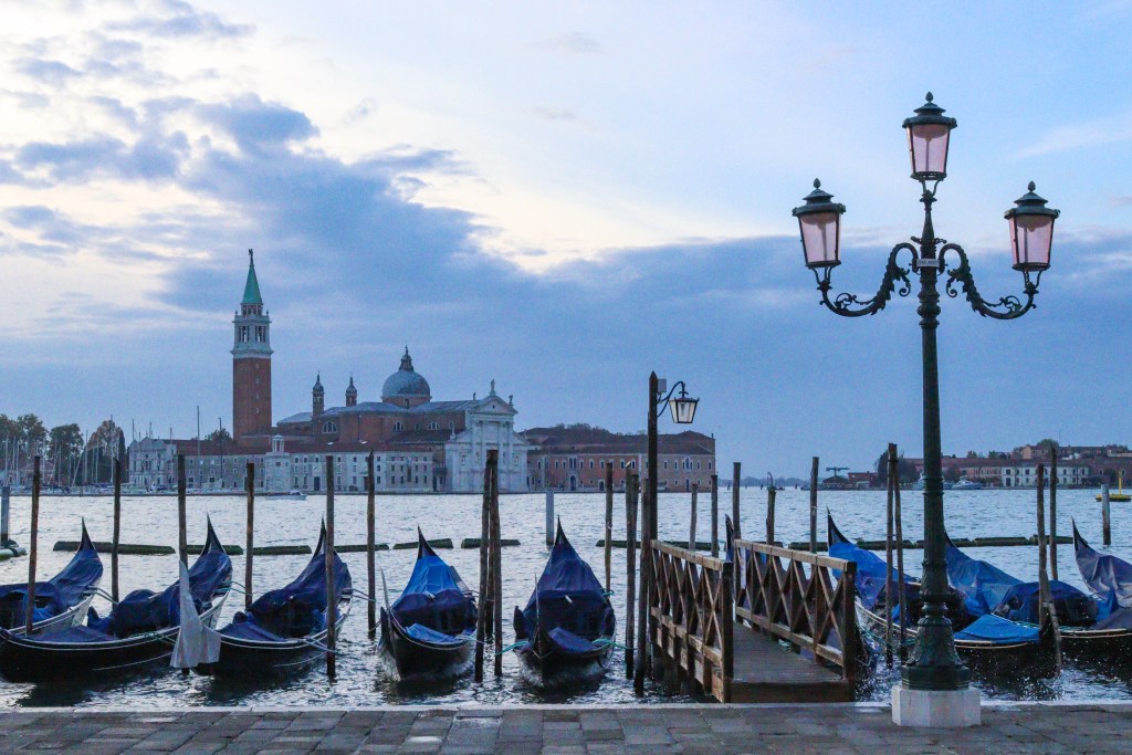 Venice: Everything You Should Know Before Your Trip Row of gondolas at Riva degli Schiavoni with lamppost and San Giorgio Maggiore at sunset in Venice.