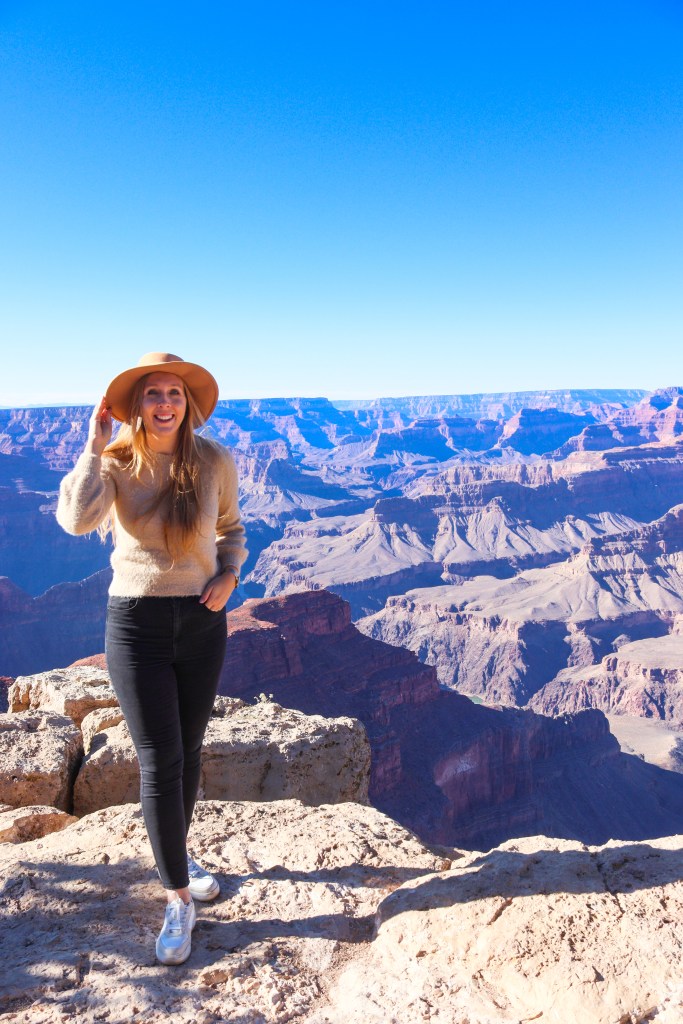 Uśmiechnięta blondynka w kapeluszu i beżowym swetrze stoi na krawędzi Grand Canyon, uśmiechając się do aparatu, z widokiem na majestatyczne formacje skalne w tle.
