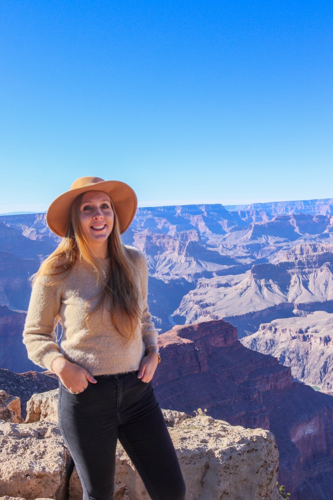 ​ Uśmiechnięta kobieta w beżowym kapeluszu i swetrze stoi na krawędzi skały nad Wielkim Kanionem, z długimi blond włosami i widokiem na kolorowe doliny w tle.
