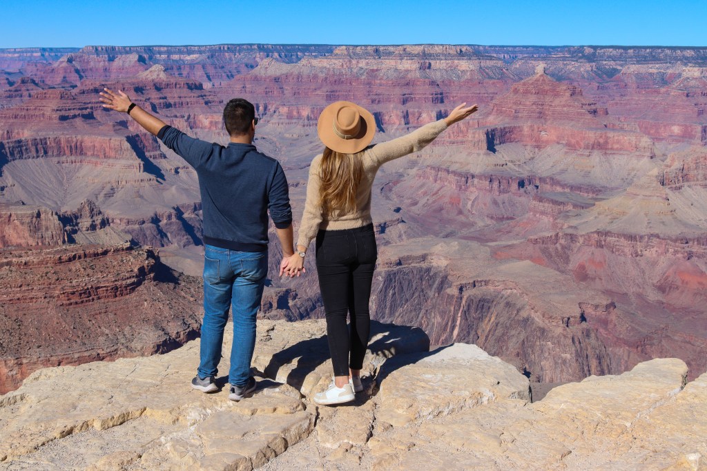 Para trzymająca się za ręce stoi tyłem na jasnej skale na krawędzi Wielkiego Kanionu, z uniesionymi ramionami i rozległą panoramą kolorowych ścian kanionu przed sobą.