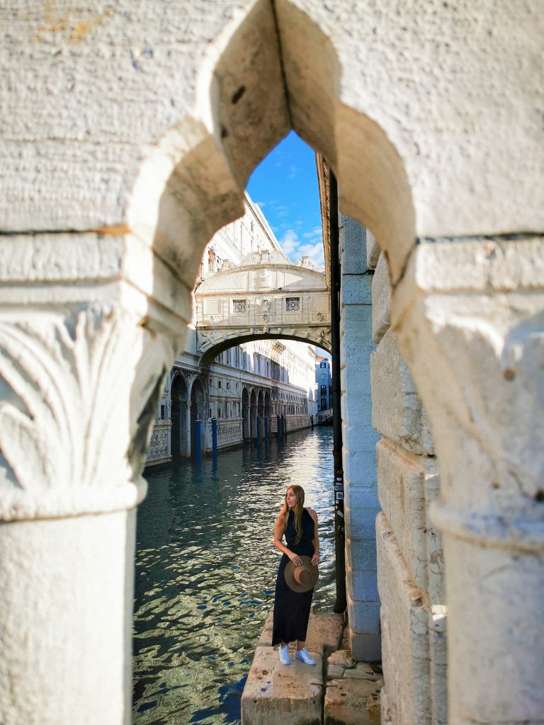 Veneza: Tudo o que deves saber antes da tua viagem Mulher junto ao canal com a Ponte dos Suspiros enquadrada por um arco de pedra