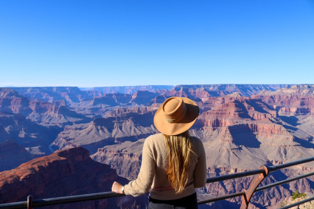 Kobieta w kapeluszu stoi tyłem, oparta o metalową barierkę na punkcie widokowym, podziwiając głębokie wąwozy i wielowarstwowe ściany Wielkiego Kanionu w ostrym, porannym słońcu.
