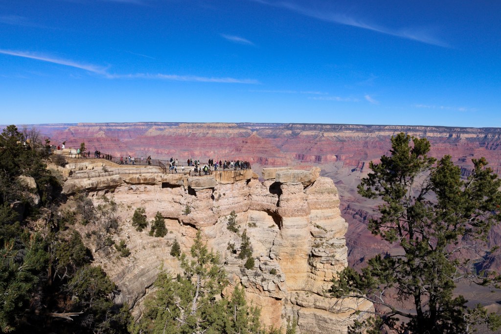 Punkt widokowy na krawędzi Wielkiego Kanionu z grupą turystów stojących za barierkami na jasnej, skalnej półce, z szeroką panoramą kolorowych ścian kanionu w tle.