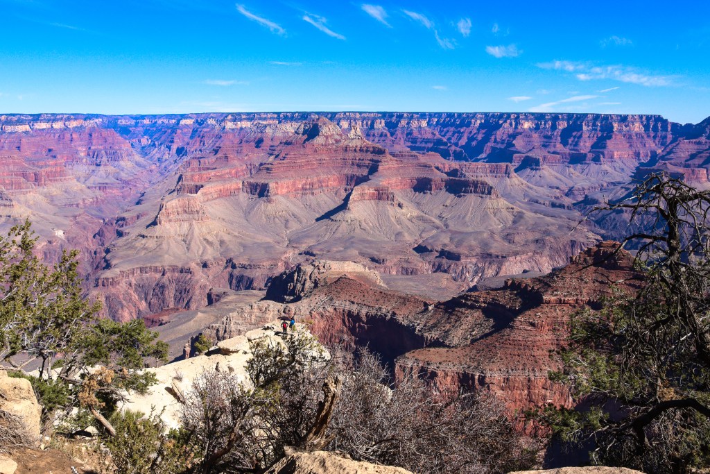 Punkt widokowy na krawędzi Wielkiego Kanionu z drzewami, osobami w oddali i rozległą panoramą warstwowych ścian w odcieniach czerwieni pod niebieskim niebem.