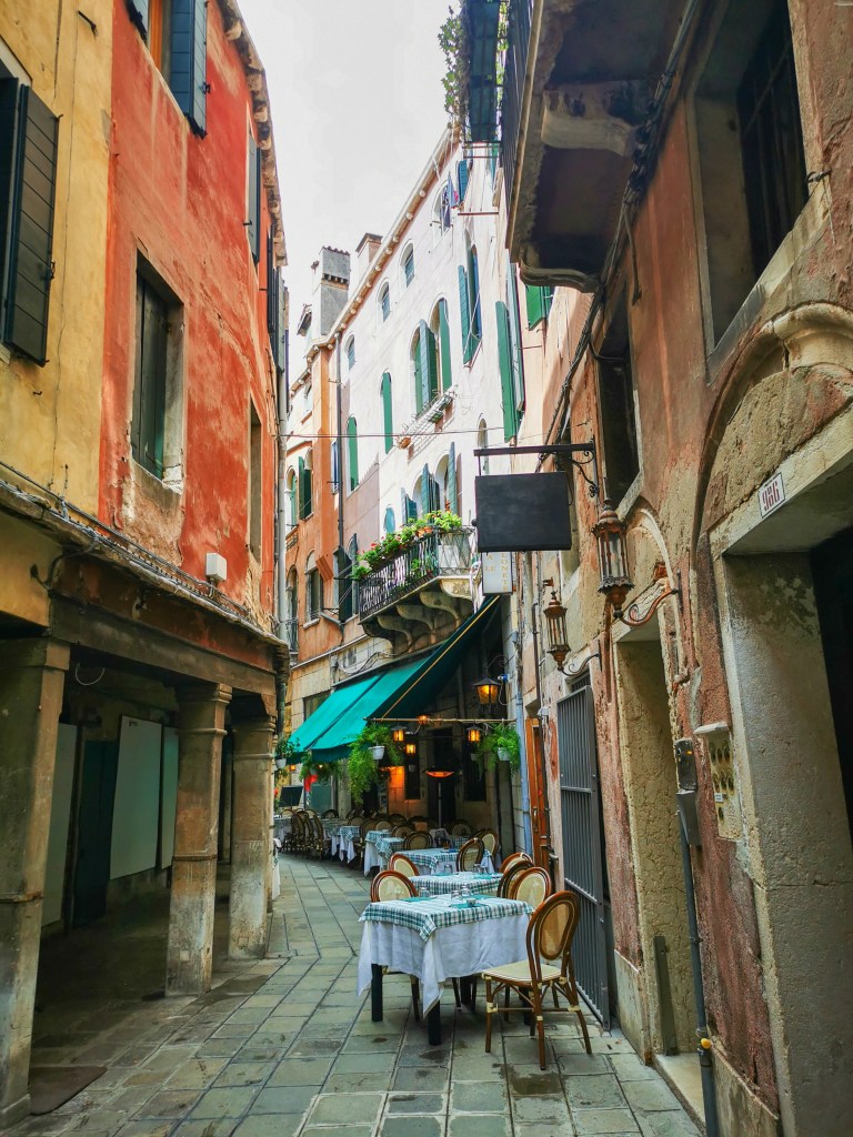Veneza: Tudo o que deves saber antes da tua viagem Beco estreito de Veneza com mesas de restaurante e casas antigas coloridas
