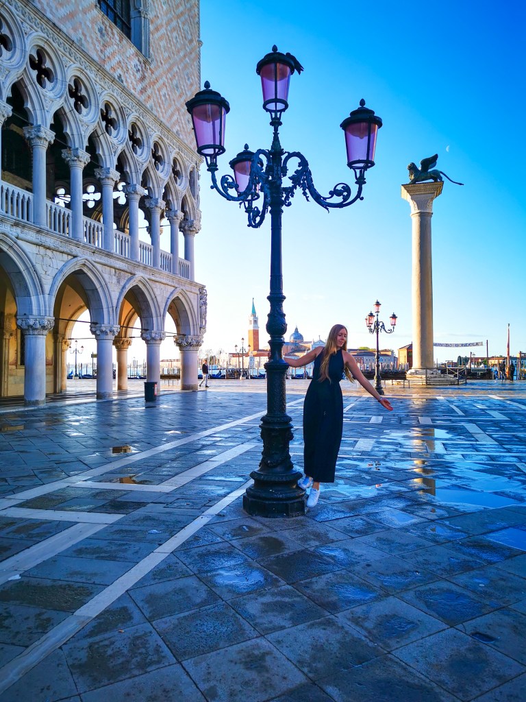 Venice: Everything You Should Know Before Your Trip Woman in black dress by a pink street lamp next to the Doge’s Palace on St Mark’s Square in Venice.