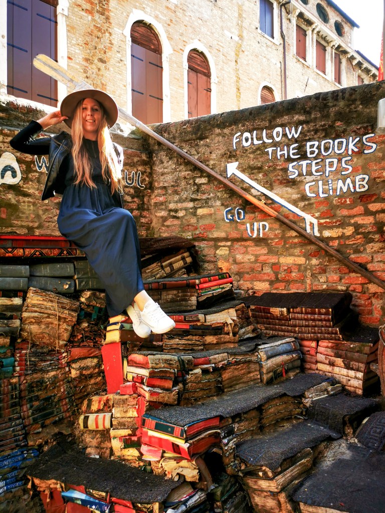 Venice: Everything You Should Know Before Your Trip Alt: Woman in hat sitting on the famous book staircase at Libreria Acqua Alta in Venice.