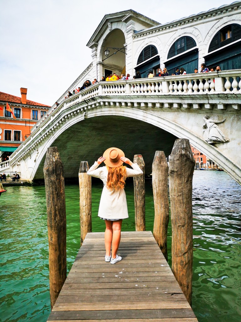 Venice: Everything You Should Know Before Your Trip Woman in hat standing on wooden pier facing the Rialto Bridge in Venice.