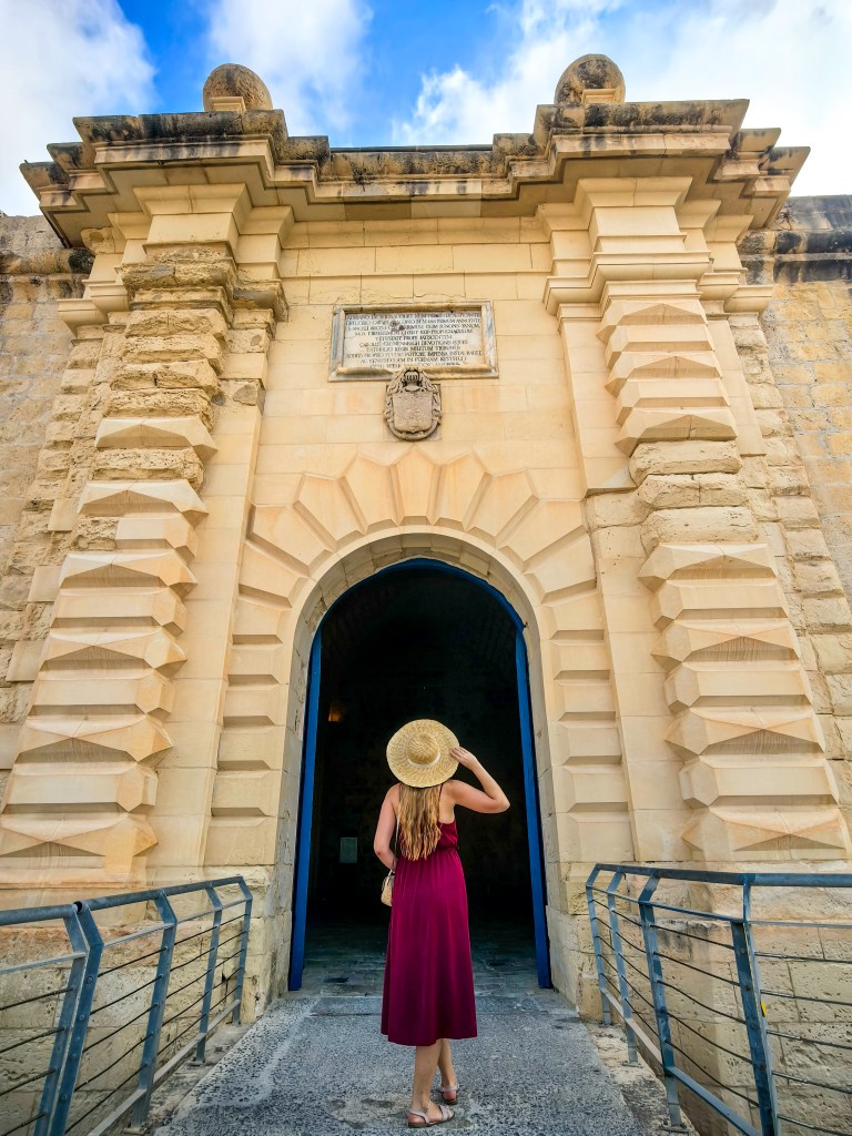 Podróżniczka w czerwonej sukience i kapeluszu stoi przed monumentalną bramą Fortu Świętego Anioła w Birgu na Malcie.