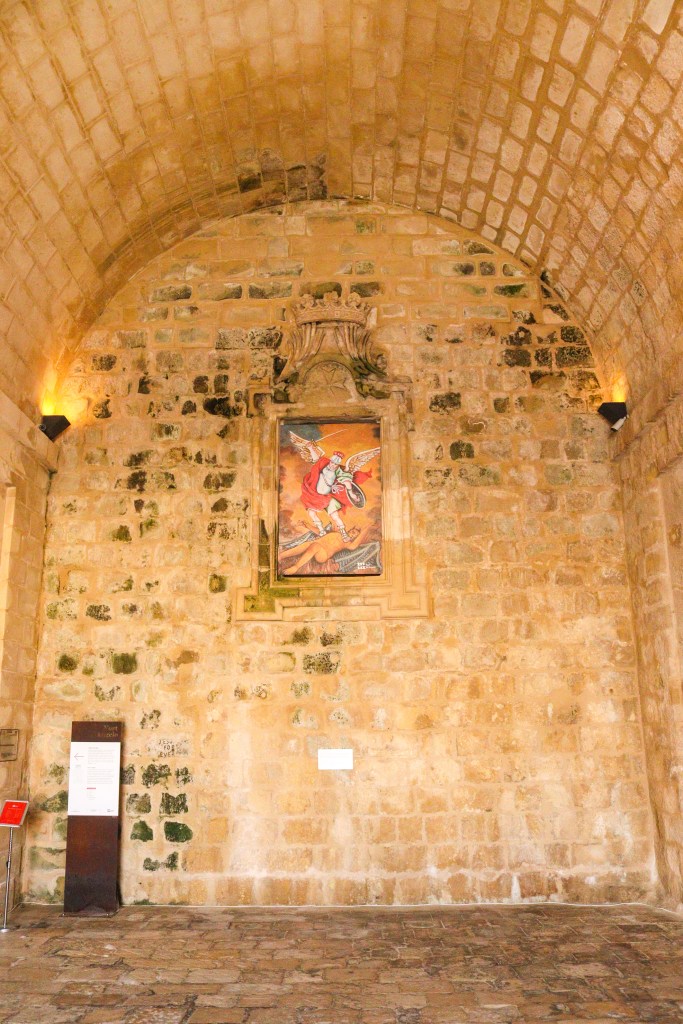 Wnętrze kamiennej sali w Forcie Świętego Anioła w Birgu z obrazem świętego na ścianie i miękkim, żółtym oświetleniem.