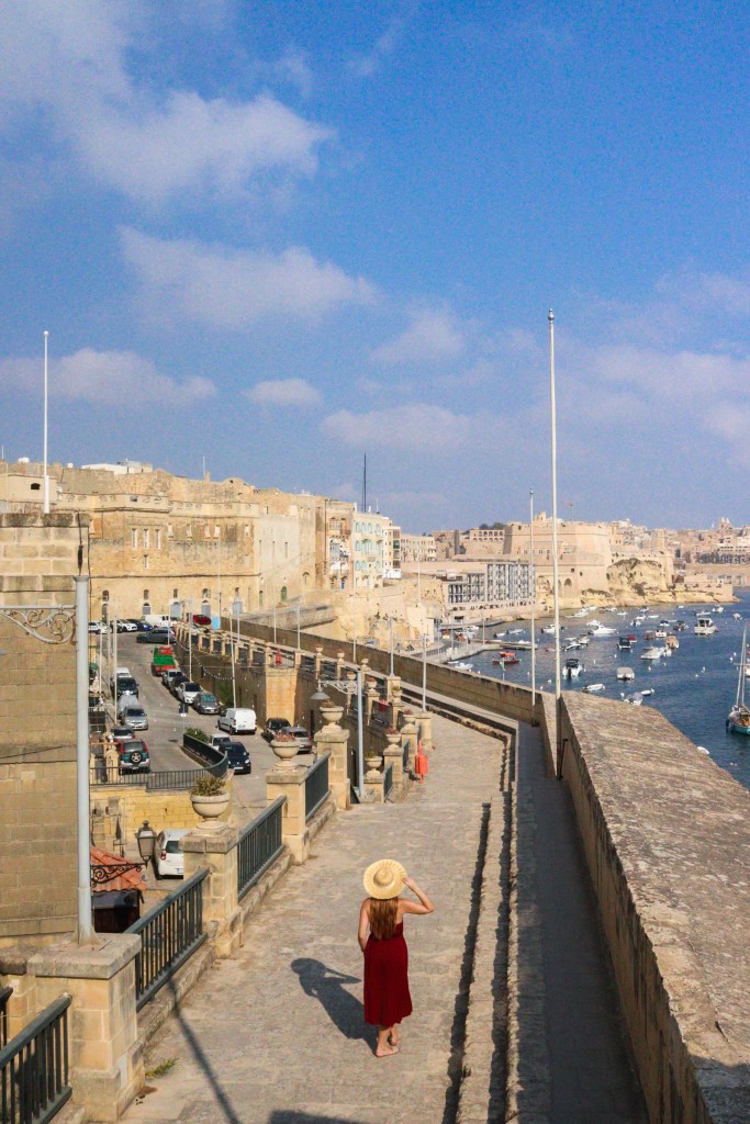 Podróżniczka w czerwonej sukience spaceruje po murach obronnych Birgu z widokiem na miasto i port.