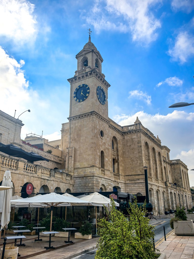 Malta Maritime Museum w Birgu z wieżą zegarową, parasolami kawiarni i stolikami na chodniku.