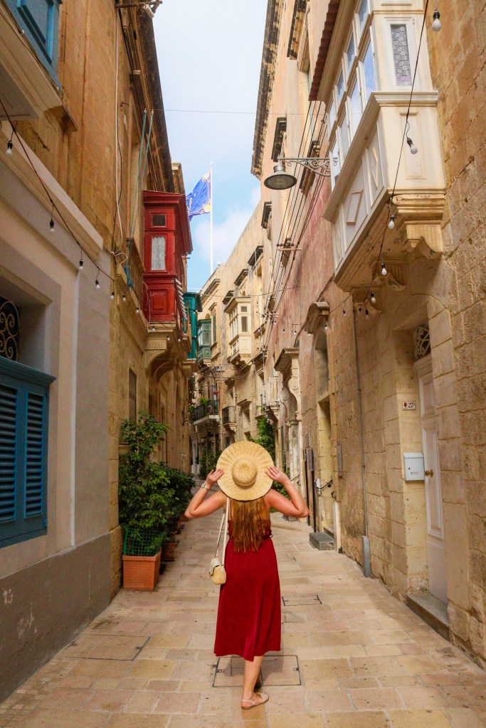 Podróżniczka w czerwonej sukience i kapeluszu idzie wąską uliczką w Birgu z kolorowymi balkonami