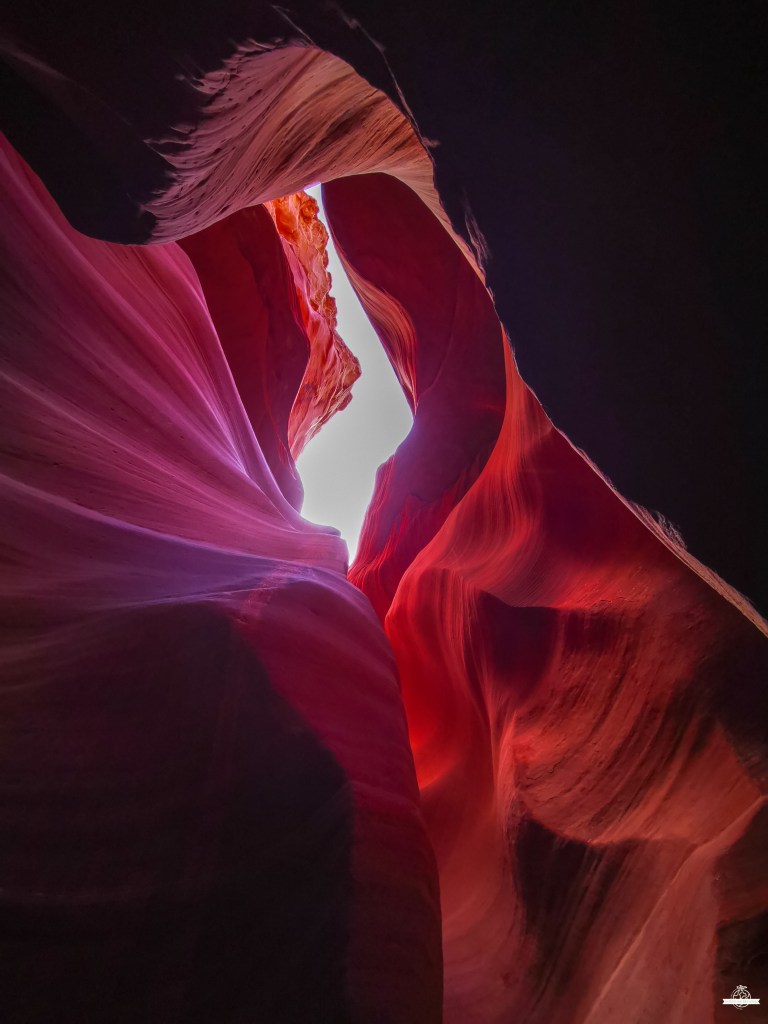 Paredes rochosas estreitas do Lower Antelope Canyon em tons magenta e vermelhos, com raios de luz branca penetrando através de fendas superiores, criando padrões luminosos nas texturas fluidas.