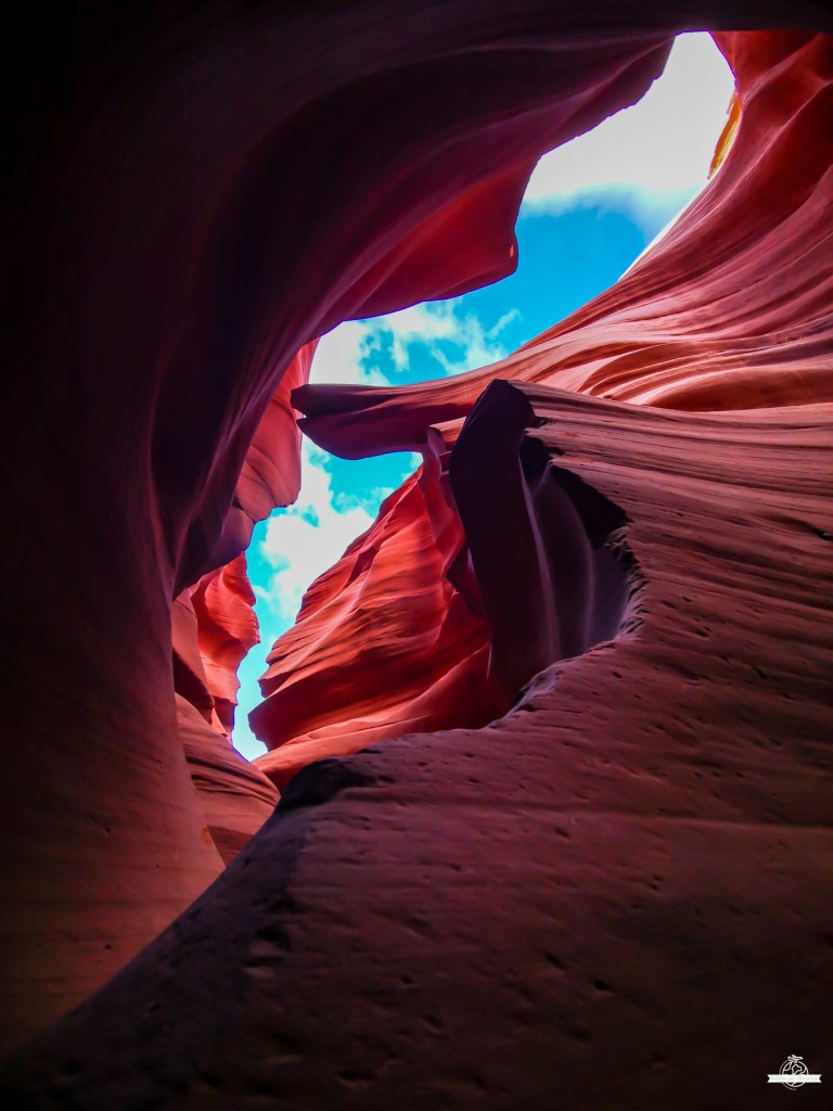 Paredes rochosas vermelhas fluidas do Lower Antelope Canyon enquadrando um céu azul com nuvens brancas através da abertura superior, com luz natural suave a iluminar as curvas dramáticas.