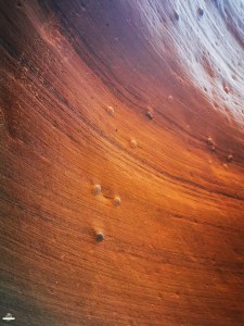 Detalhe macro das formações rochosas fluidas do Lower Antelope Canyon, com texturas onduladas em tons de vermelho, laranja e roxo, salpicadas por partículas de areia em suspensão.