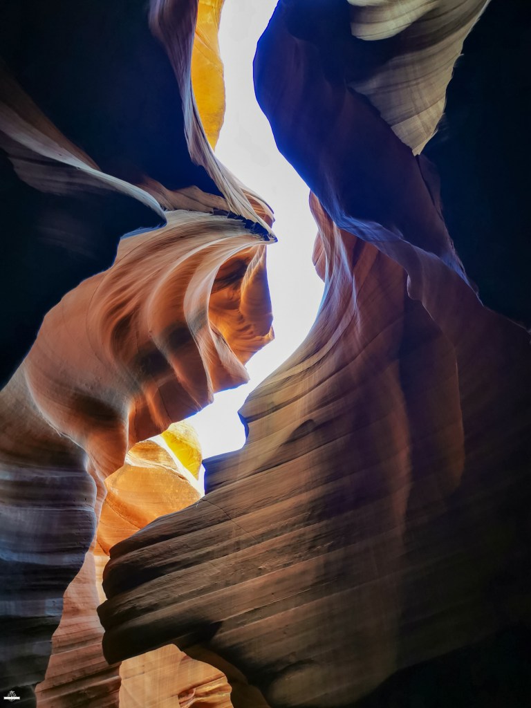 Formações rochosas fluidas do Lower Antelope Canyon iluminadas por um feixe de luz solar intensa em tons de amarelo e laranja, contrastando com sombras escuras nas paredes avermelhadas e texturas onduladas.