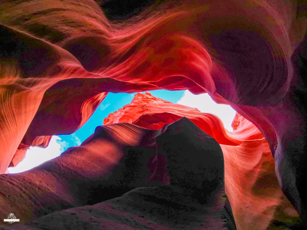 Formações rochosas dramáticas do Lower Antelope Canyon em tons vermelhos profundos, enquadrando um pedaço de céu azul através da abertura superior, com luz natural a realçar as curvas fluidas das paredes arenosas.