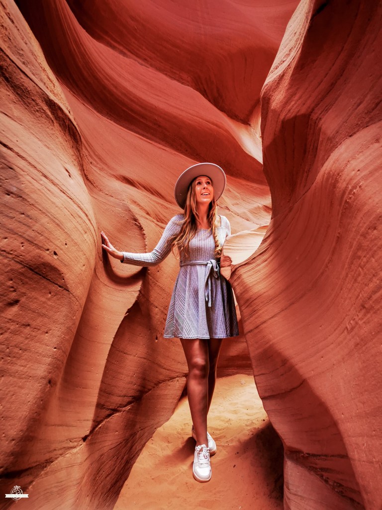 Mulher com chapéu e vestido azul claro apoiada numa rocha no Lower Antelope Canyon, rodeada por paredes rochosas fluidas em tons vermelhos e laranja.