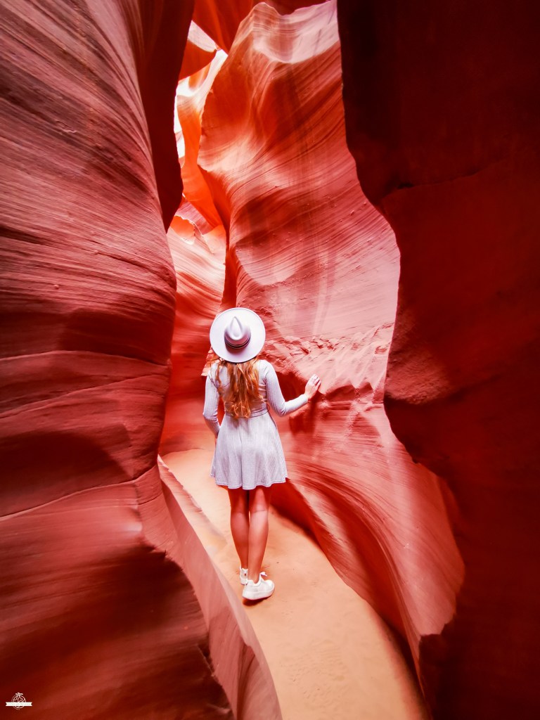 Mulher vista de costas com chapéu e vestido azul a caminhar num estreito corredor do Lower Antelope Canyon, ladeado por paredes rochosas onduladas em tons vermelhos vibrantes.