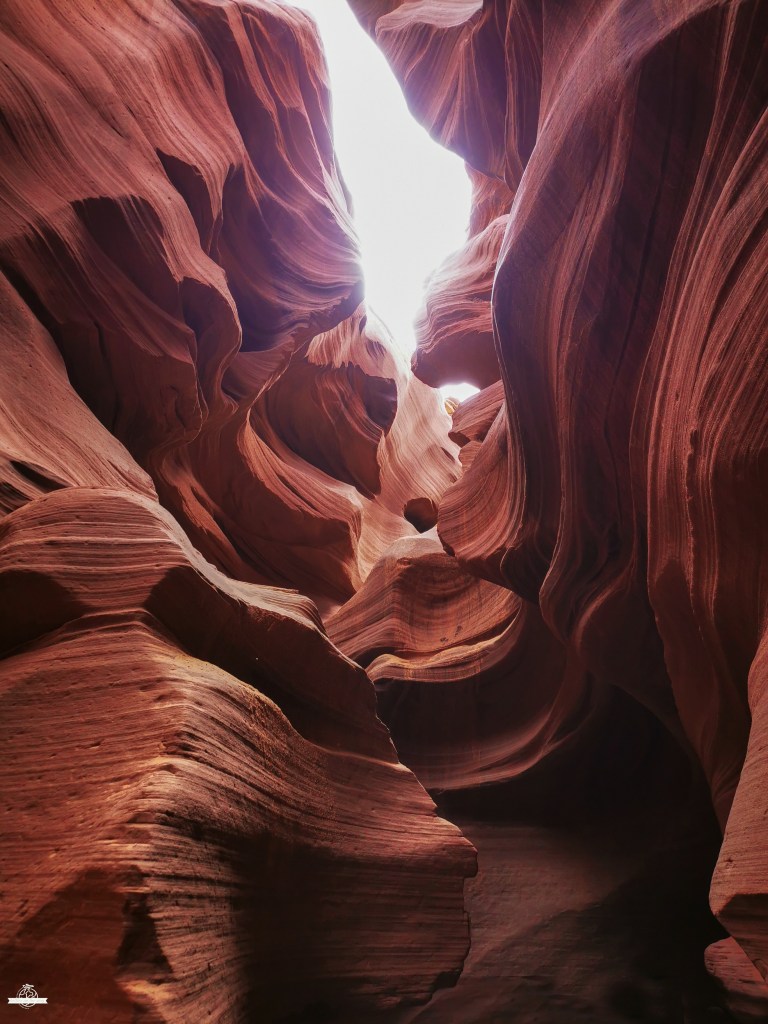 Paredes rochosas fluidas do Lower Antelope Canyon em tons laranja e vermelhos, iluminadas por um feixe de luz solar brilhante que cria contrastes dramáticos nas curvas arenosas.