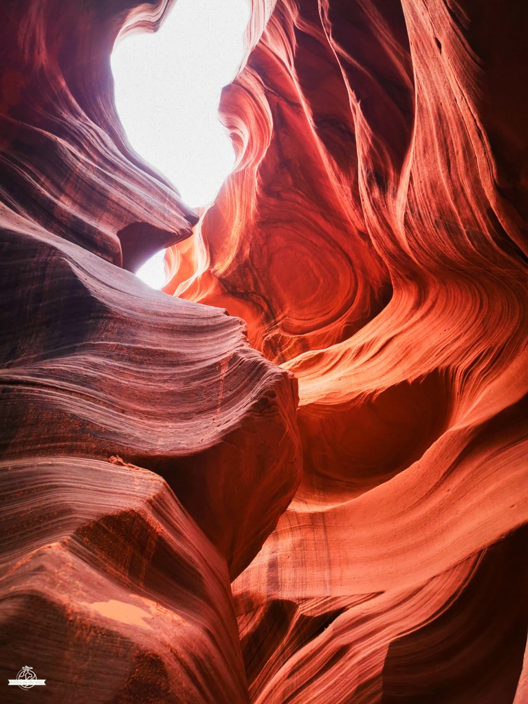 Paredes rochosas fluidas do Lower Antelope Canyon em tons vermelhos e laranja, iluminadas por um feixe de luz branca intensa que penetra do topo, realçando padrões circulares e curvas dramáticas nas texturas arenosas.