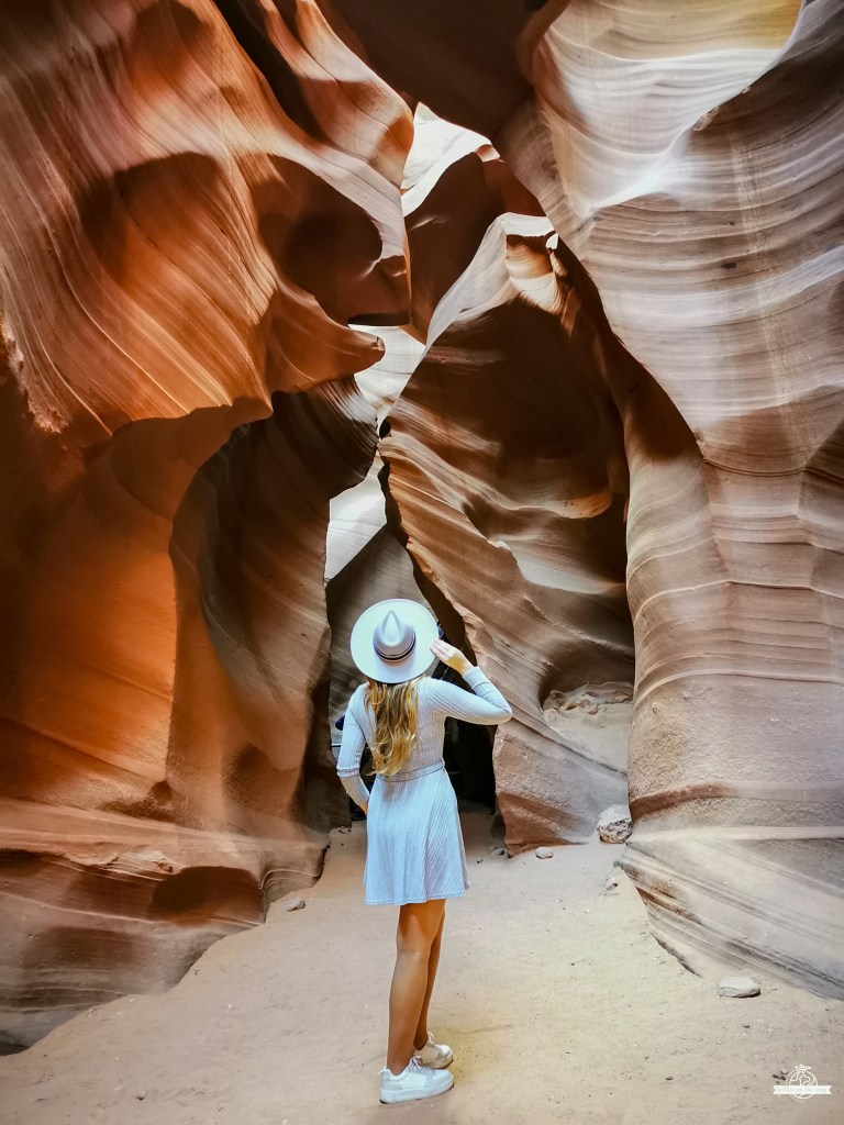 Mulher loira vista de costas com chapéu e vestido azuis claro a caminhar para a entrada estreita do Lower Antelope Canyon, ladeada por paredes rochosas fluidas em tons vermelhos e laranja.