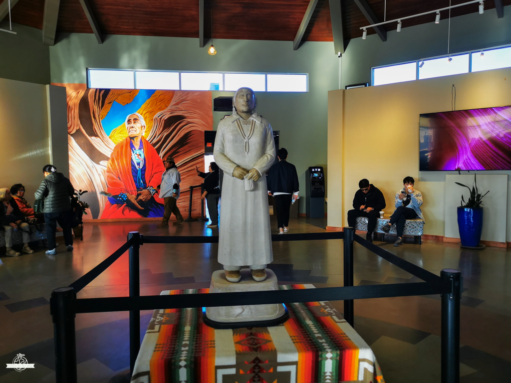Estátua de um nativo Navajo em vestes tradicionais brancas com detalhes coloridos, de pé numa base com manta listrada multicolor, num espaço de visitantes do Lower Antelope Canyon rodeado por murais abstratos vermelhos e turistas.