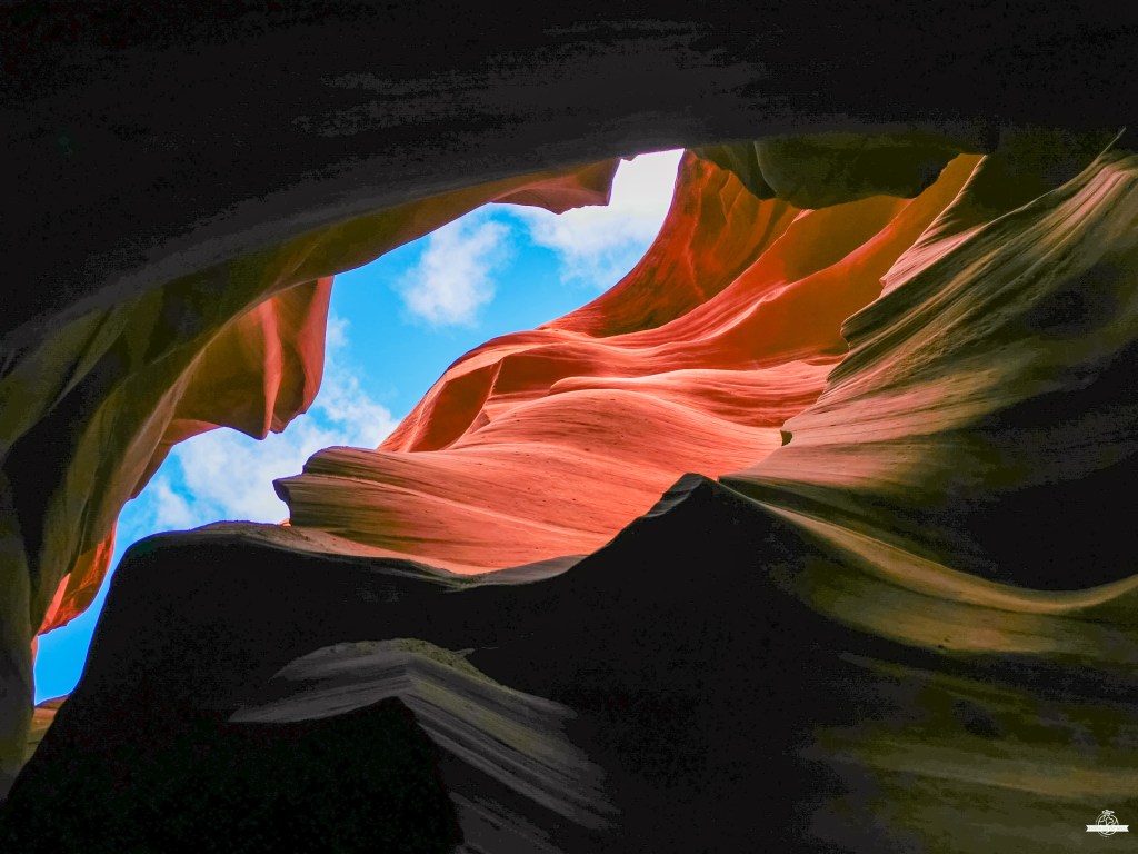 Paredes rochosas fluidas em tons vermelhos e laranja do Lower Antelope Canyon enquadrando um céu azul com nuvens brancas através da abertura superior, criando um contraste dramático entre as sombras escuras e a luz natural.