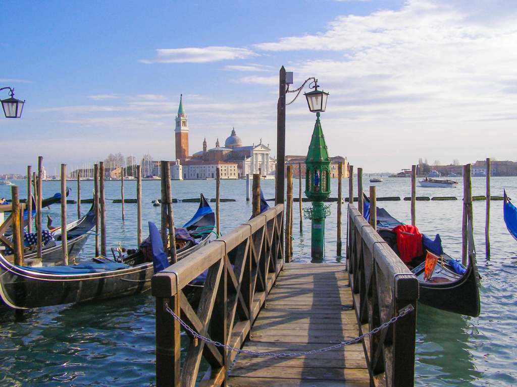 Wenecja – jak się poruszać po Mieście Dożów? Drewniany pomost z gondolami na Riva degli Schiavoni w Wenecji i widokiem na wyspę San Giorgio Maggiore z kościołem i dzwonnicą.