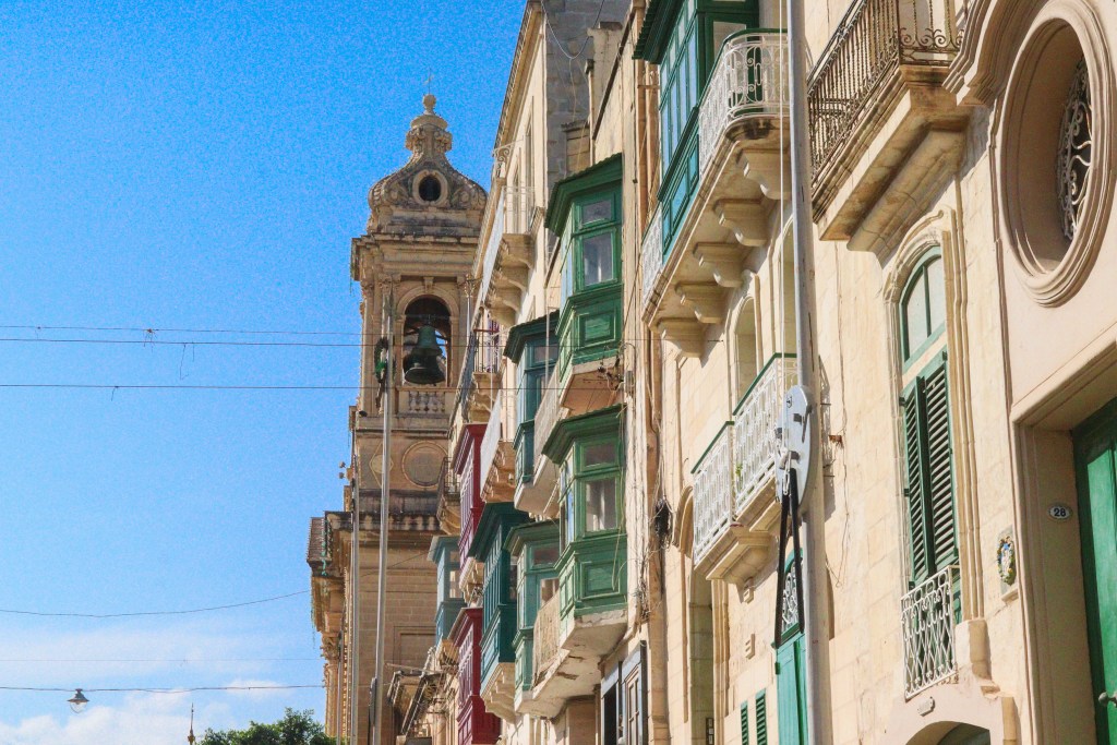 Ulica w Isli z kolorowymi zabudowanymi balkonami i wysoką dzwonnicą kościoła na tle błękitnego nieba.