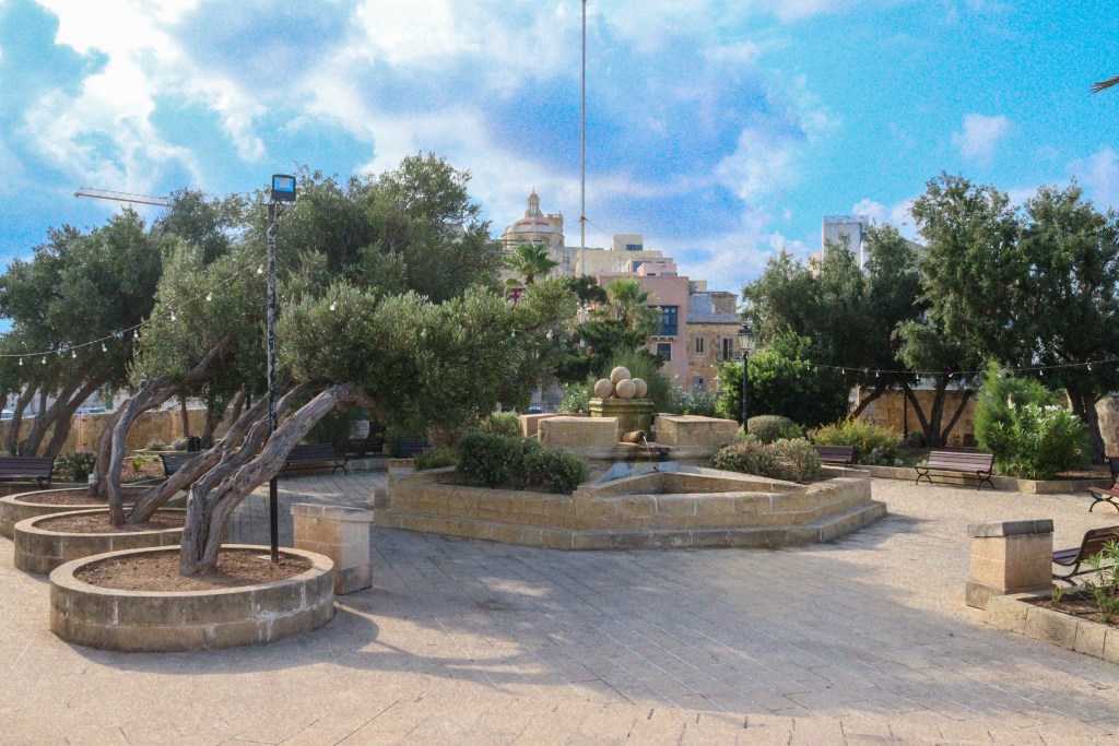 Uroczy Ogród Guardjola w Isli z fontanną, drzewami oliwnymi i ławkami pośród piaskowych murków.