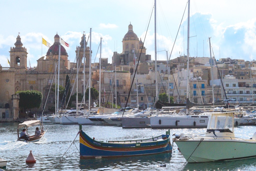 Kolorowe łódki i jachty w porcie Isli z barokowym kościołem i piaskowymi kamienicami Birgu w tle.