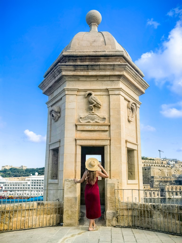 Podróżniczka w czerwonej sukience przy ozdobnej wieży strażniczej Il-gardjola w Guardjola Gardens w Isli z widokiem na morze i Vallettę.