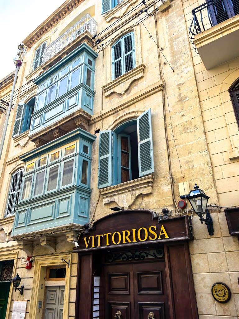 Niebieskie i drewniane balkony nad wejściem do kawiarni w Vittoriosa w Birgu, z lampami i kwiatami.