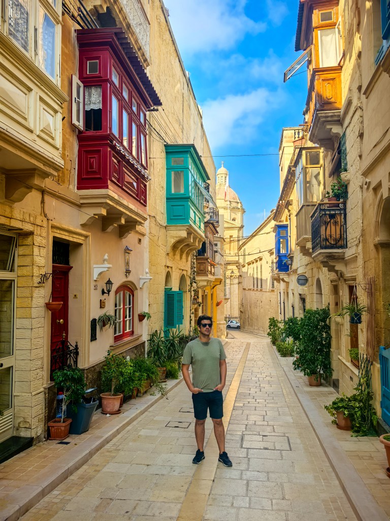 Kolorowa uliczka w Vittoriosa z czerwonymi i zielonymi balkonami, podróżnik pozuje na tle kościoła.