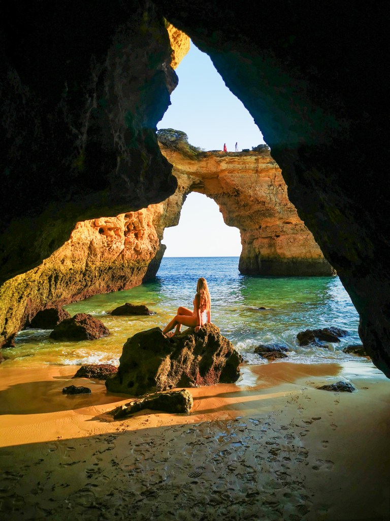 Our Top 7 Beaches in the Algarve Woman sitting in a cave near Praia de Albandeira with Arco de Albandeira in the background