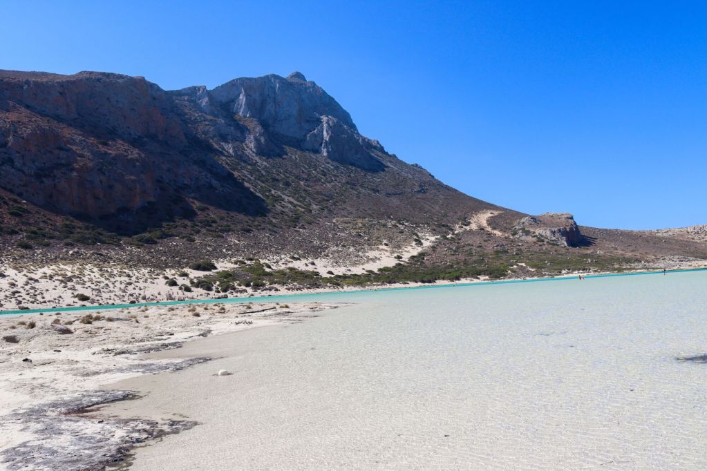 Wide sandy shore of Balos Lagoon with the massive cliffs of the Gramvousa Peninsula in the background.