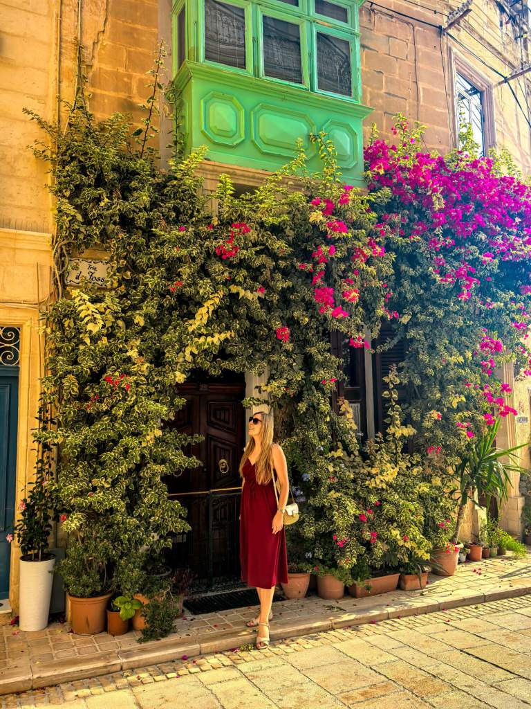 A Guide to Malta’s Three Cities: Discover Birgu, Isla and Bormla A woman in a red dress standing in front of a historic doorway covered in blooming pink bougainvillea on Triq il-Foss street in Birgu.