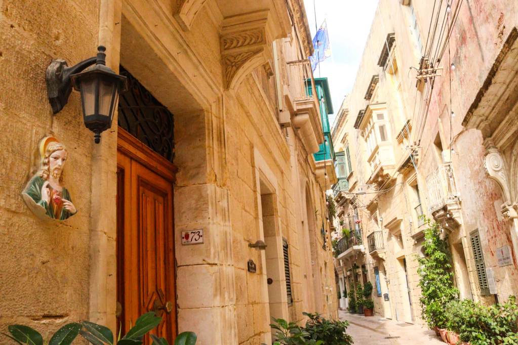 A Guide to Malta’s Three Cities: Discover Birgu, Isla and Bormla A narrow charming street in Birgu with a religious shrine on the wall and traditional limestone architecture.