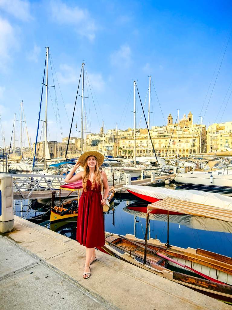 A Guide to Malta’s Three Cities: Discover Birgu, Isla and Bormla A woman in a red dress and straw hat standing on a wooden dock at the Birgu marina surrounded by yachts.
