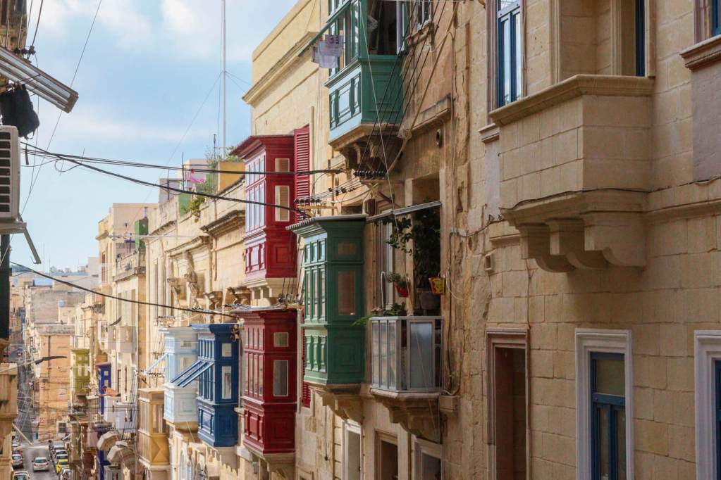 A Guide to Malta’s Three Cities: Discover Birgu, Isla and Bormla A narrow limestone street in Isla (Senglea) featuring several storeys of traditional red, green, and blue wooden balconies.