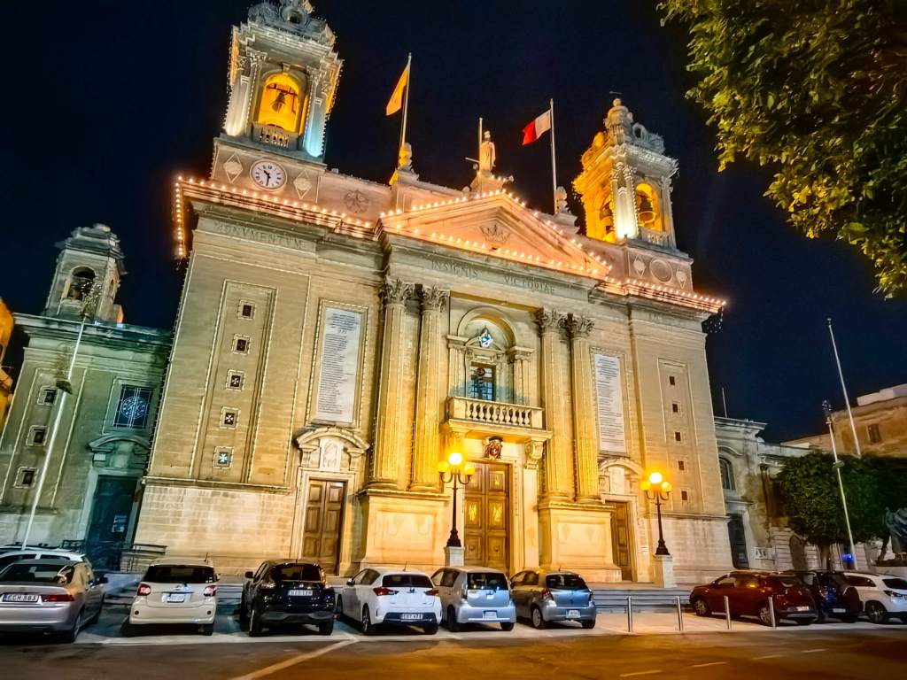 A Guide to Malta’s Three Cities: Discover Birgu, Isla and Bormla The illuminated facade of the Basilica of the Nativity of Mary in Senglea at night with parked cars in the foreground.