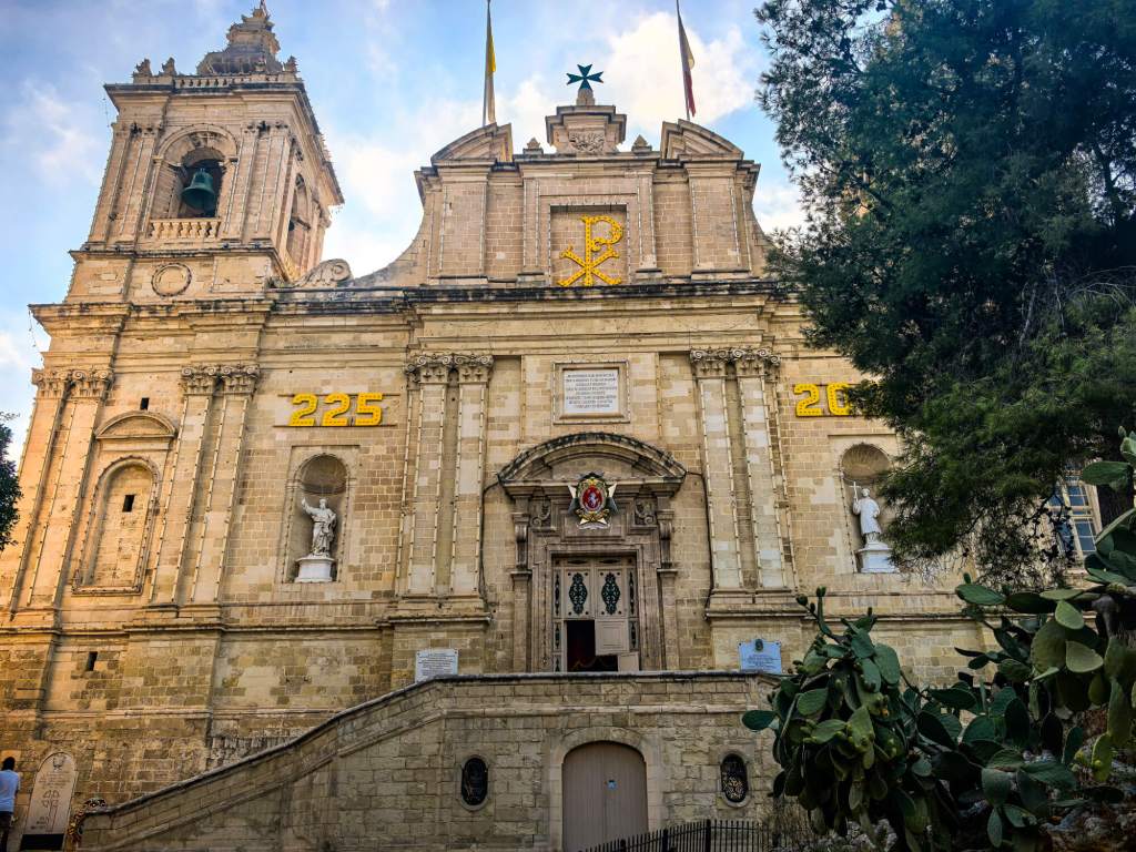 A Guide to Malta’s Three Cities: Discover Birgu, Isla and Bormla The detailed Baroque facade of St Lawrence Collegiate Church in Birgu with a large prickly pear cactus in the foreground.